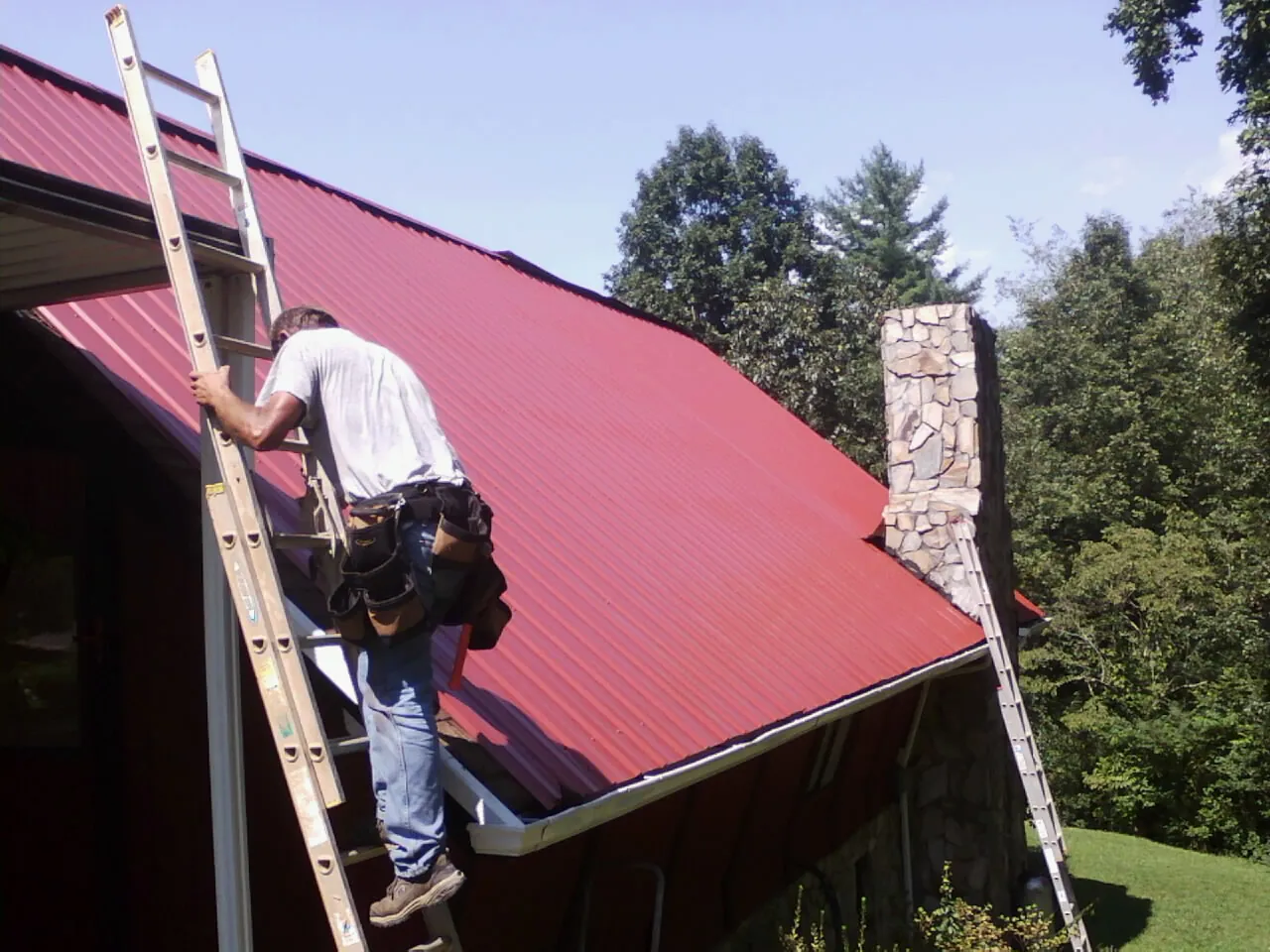 Roofer on a ladder beside a red metal roof and stone chimney — Soffit & Fascia Repair by Roofing Brothers in Platteville
